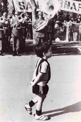 Primer plano de niño, caminando tranquilamente por delante de una banda instrumental de militares