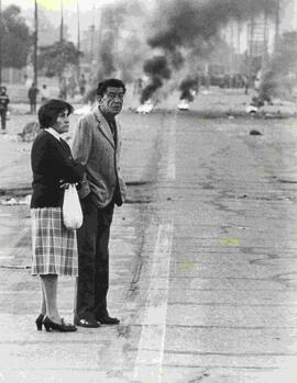 Dos adultos observan a la cámara mientras de fondo se observan barricadas